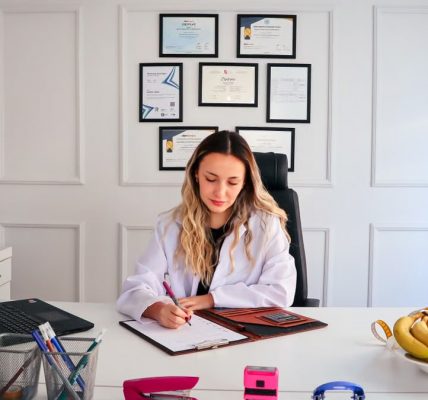 Nutritionist sits at a modern office desk writing, with fruits and diplomas displayed, emphasizing health and professionalism.